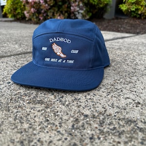 Könnte beinhalten: Eine marineblaue 5-Panel-Kappe mit einem weißen gestickten Logo, auf dem "Dadbod Run Club" und "One Mile at a Time" steht. Das Logo zeigt auch einen stilisierten orangen Laufschuh mit Flügeln.