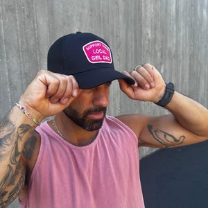 May include: A black mesh trucker hat with a pink patch that says "Support Your Local Girl Dad".