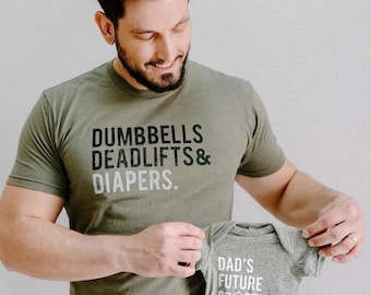 May include: A green t-shirt with the text "Dumbbells Deadlifts & Diapers." A person is holding a gray baby onesie with the text "Dad's Future Spotter."