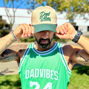 May include: A tan and green baseball cap with the text "Dad Vibes" embroidered in green. The cap is being held by a person wearing a green basketball jersey with the text "DADVIBES" and the number "24" on the front.
