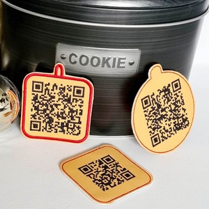 Könnte beinhalten: Vier gelbe und rote Weihnachtsbaumschmuck-förmige Geschenkanhänger mit schwarzen und weißen QR-Codes. Die Anhänger liegen auf einer schwarzen Keksdose mit dem Wort "COOKIE" auf der Vorderseite. Im Hintergrund sind ein goldener Stern und zwei silberne Kugeln zu sehen.