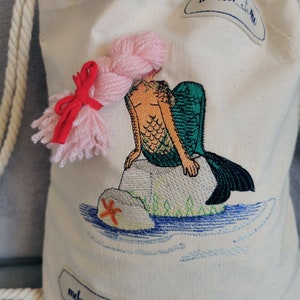 May include: A white canvas tote bag with an embroidered mermaid design. The mermaid has pink hair, a green tail, and is sitting on a rock with a starfish. The bag has a rope handle.