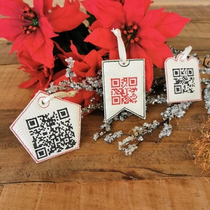 Könnte beinhalten: Drei weiße Geschenkanhänger mit roten und schwarzen gestickten QR-Codes. Die Anhänger hängen an einem Zweig einer Weihnachtsstern-Pflanze mit silbernem Glitzer.