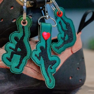 Könnte beinhalten: Drei grüne Leder-Schlüsselanhänger mit schwarzen Silhouette-Figuren und roten Herz-Akzenten. Die Schlüsselanhänger sind an einem Paar schwarzer und rosa Kletterschuhe befestigt.