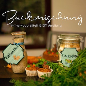 May include: Two glass jars with lids, filled with baking mix, labeled "Rublimuffins" and "Rublikuchen". The jars are on a wooden table with carrot cake cupcakes, carrots, and green foliage.
