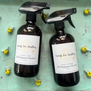 May include: Two amber glass spray bottles with white labels. The labels feature a honeycomb design and the text "Honey I'm Healthy" with the scent and size listed below. The bottles are on a teal surface with yellow bee decorations.