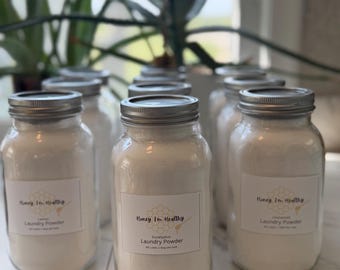 Puede incluir: Varios frascos de vidrio transparente llenos de detergente en polvo blanco, cada uno con una tapa de metal plateado. Los frascos tienen etiquetas blancas con el texto Honey I'm Healthy, un gráfico de panal y el aroma específico.