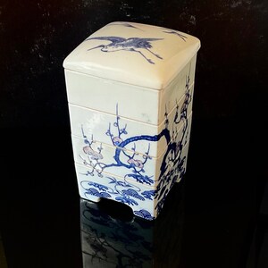 Op de afbeelding: Een witte, geglazuurde doos met vier verdiepingen, versierd met blauwe en roze bloemen en een kraanvogel op de bovenste verdieping. De doos is in traditionele Japanse stijl.