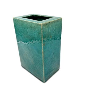 May include: A rectangular ceramic vase with a glossy teal glaze. The surface features a subtle crackle pattern and a distinct horizontal white line running across the front face. The vase has a flat top opening and sharp, defined vertical edges.
