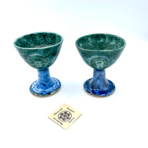 May include: Two ceramic goblets in teal and blue. The bowls are teal, and the stems and bases are blue. Each goblet has a circular emblem with the text "STONEHENGE". A small card with text is in the foreground.