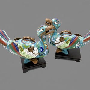 Pode incluir: Três recipientes decorativos em forma de pássaro com intrincados desenhos de esmalte. Os pássaros apresentam padrões turquesa, vermelhos, azuis e verdes, com bicos e alças de cor bronze. Cada um assenta numa base quadrada preta.