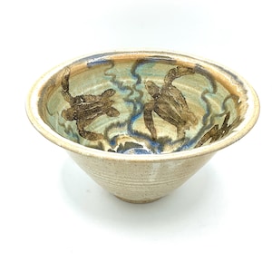 Może przedstawiać: Beżowa ceramiczna miska z szerokim brzegiem i lekko zwężającym się kształtem. Wnętrze zdobi jasnoniebieski i brązowy wzór z dwoma brązowymi żółwiami morskimi. Misa ma teksturowaną powierzchnię zewnętrzną.