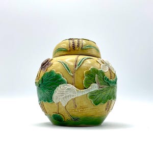 May include: A yellow ceramic jar with a lid, featuring a white crane, green lily pads, and white flowers. The jar has a round shape and is decorated with raised relief details. The lid has a brown and green design. The jar is approximately 8 inches tall.