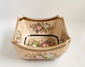 May include: A square ceramic bowl with a cream-colored base and gold trim. The bowl is decorated with a floral pattern in shades of pink, red, green, and white.
