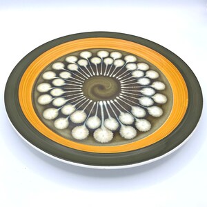 Peut inclure: Une grande assiette ronde en céramique avec un motif brun, jaune et blanc. Le centre de l'assiette présente un motif tourbillonnant de points blancs sur un fond brun. Le bord extérieur de l'assiette a une bande jaune avec une bordure brune.