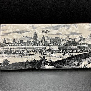 May include: A black and white engraving of the city of Giessen, Germany. The engraving shows the city's skyline, including a church with a tall spire, and a surrounding landscape.