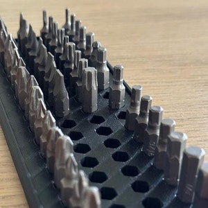 May include: A black plastic organizer with a honeycomb pattern holds a variety of small metal screwdriver bits.