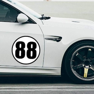 May include: White sports car with a black and white racing number 88 decal on the side. The car has black rims and a yellow accent on the brake caliper. The background is a parking lot with light poles.