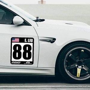 May include: White car with a black and white racing number sticker on the side. The sticker has the number 88 and the text "E. LIU" on it. The sticker also has a small American flag.