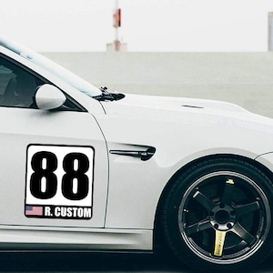 May include: A white car with a black and white sticker that has the number 88 in a bold font. Below the number is the text "R. CUSTOM" and a small American flag. The car is parked on a paved surface.