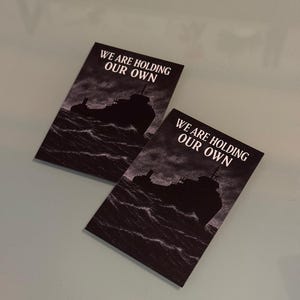 Peut inclure: Deux cartes rectangulaires pr&eacute;sentant une illustration en noir et blanc d'un navire naviguant sur des vagues sombres et agit&eacute;es. Le texte WE ARE HOLDING OUR OWN est imprim&eacute; en lettres capitales blanches en haut de chaque carte sur un ciel sombre.