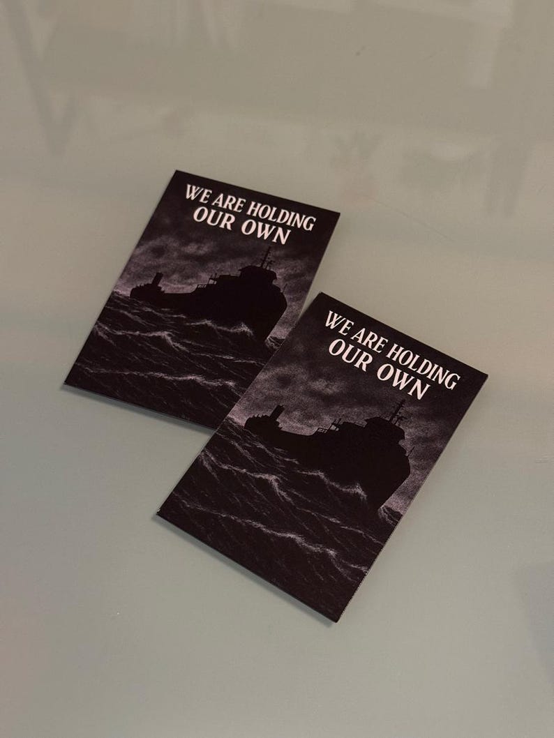 Peut inclure: Deux cartes rectangulaires pr&eacute;sentant une illustration en noir et blanc d'un navire naviguant sur des vagues sombres et agit&eacute;es. Le texte WE ARE HOLDING OUR OWN est imprim&eacute; en lettres capitales blanches en haut de chaque carte sur un ciel sombre.