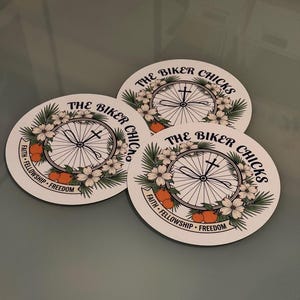 Peut inclure: Trois autocollants circulaires blancs pr&eacute;sentant un dessin au trait noir d'une roue de v&eacute;lo centr&eacute;e avec une croix. Le motif inclut des fleurs blanches, des feuilles vertes et des fruits orange. Le texte indique THE BIKER CHICKS et FAITH FELLOWSHIP FREEDOM.