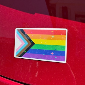 May include: A rectangular sticker featuring the Progress Pride flag. The flag design includes the traditional rainbow stripes, along with a chevron incorporating colors representing transgender, marginalized, and people of color. The sticker has a white border.
