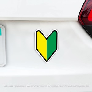 May include: A yellow and green beginner driver sticker in a V-shape, with a black border. The sticker is affixed to a white car surface. The text at the bottom reads: *NOT SCALED TO SIZE, COLORS MAY DISPLAY DIFFERENTLY ON YOUR MONITOR THAN WHAT IS ACTUALLY PRINTED*