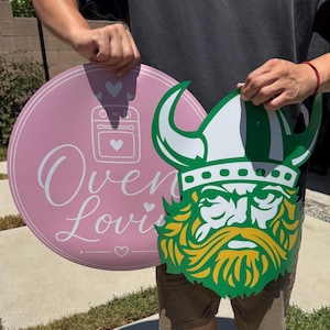 Peut inclure: Un panneau circulaire rose avec une ic&ocirc;ne de four blanche, des c&oelig;urs et le texte "Oven Lovin" &agrave; c&ocirc;t&eacute; d'un panneau d&eacute;coup&eacute; repr&eacute;sentant une t&ecirc;te de Viking avec un casque blanc, une bordure verte et une barbe jaune, en mat&eacute;riau rigide.