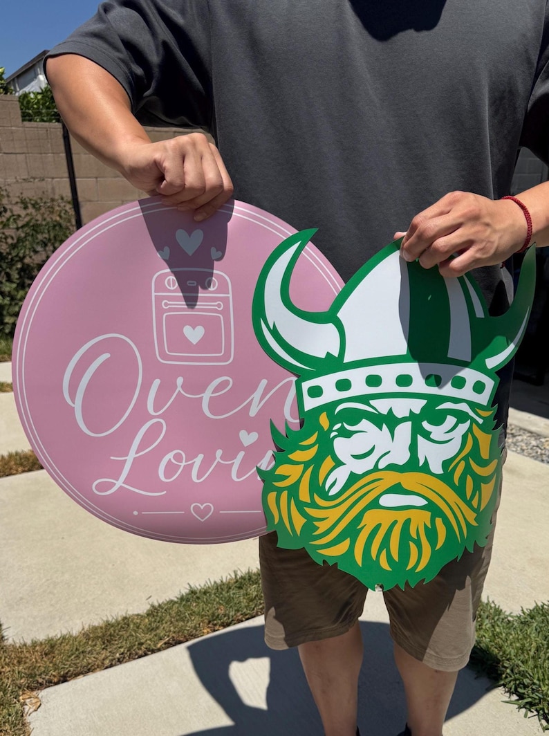 Peut inclure: Un panneau circulaire rose avec une ic&ocirc;ne de four blanche, des c&oelig;urs et le texte "Oven Lovin" &agrave; c&ocirc;t&eacute; d'un panneau d&eacute;coup&eacute; repr&eacute;sentant une t&ecirc;te de Viking avec un casque blanc, une bordure verte et une barbe jaune, en mat&eacute;riau rigide.