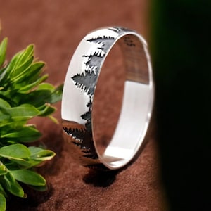 May include: A silver band ring with a design of a row of pine trees etched into the metal.