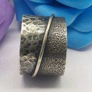 Puede incluir: Un anillo de banda ancha de plata con un acabado martillado texturizado y una banda de plata lisa envuelta alrededor.