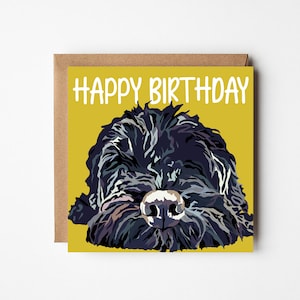 May include: A square greeting card with a mustard yellow background featuring a stylised illustration of a shaggy black dog face. The text HAPPY BIRTHDAY is printed in white capital letters at the top. A brown envelope is positioned behind the card.