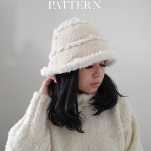 May include: A white knitted bucket hat with a fluffy white trim. The hat is being worn by a person with long black hair. The text "Vivienne Bucket Hat Knitting Pattern" is at the top of the image.