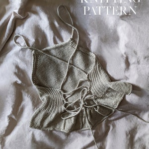 Puede incluir: Un top tejido de color beige claro con espalda cruzada y cierre de lazo. El top tiene tirantes finos y un dobladillo con volantes. Las palabras "KNITTING PATTERN" están impresas en blanco.