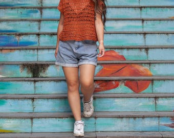 May include: An orange crochet top with short sleeves and a round neckline. The top is paired with denim shorts and white sneakers. The text "Hollyberry Tee KNITTING PATTERN" is at the bottom of the image.