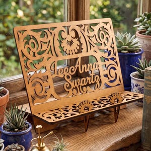 May include: A laser-cut wooden book stand featuring intricate scrollwork patterns and the name LeeAnna Swartz engraved in the centre. The stand includes a horizontal shelf with two decorative supports and a slanted back panel for holding books.