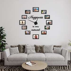 Op de afbeelding: Een zwarte fotolijstklok met de woorden "Happiness in every moment" op een witte muur. De klok is omringd door 12 kleinere zwarte fotolijsten, die elk een foto van een stel bevatten.