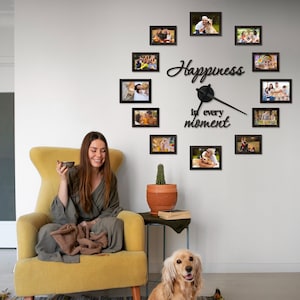 Peut inclure: Horloge murale décorative avec des cadres photo noirs disposés en cercle. Le cadran de l'horloge affiche le texte "Happiness in every moment". Une femme est assise dans un fauteuil jaune, avec un chien.