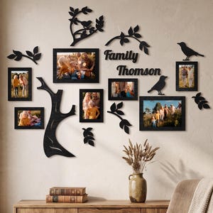 Könnte beinhalten: Ein schwarzes Wand-Fotorahmen-Set mit Baumstamm, Ästen und Vogelsilhouetten. Das Arrangement umfasst fünf rechteckige Rahmen mit Familienfotos sowie den schwarzen, ausgeschnittenen Schriftzug Family Thomson.