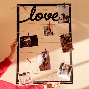 May include: A black rectangular photo display with the word "love" in cursive. Photos are clipped to twine with small pink and red clothespins. The display is held by a person wearing a red sweater.