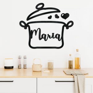 Puede incluir: Arte de pared de metal negro con forma de olla con tapa, asas y corazones. El nombre "Maria" está recortado dentro de la olla. Montado en una pared blanca sobre una encimera de madera con accesorios de cocina.