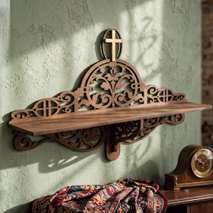 Puede incluir: Un estante de madera montado en la pared con intrincados patrones florales y de volutas cortados con láser. El diseño incluye un motivo circular central coronado por una pequeña cruz dorada vertical. El estante tiene una superficie plana.