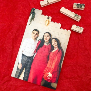 May include: A photo puzzle featuring a family of three. The photo shows a man and two women standing together. The man is wearing a white shirt and dark pants. The women are wearing red dresses. The puzzle is made up of white plastic pieces.