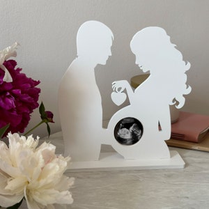 May include: White silhouette of a couple holding a heart and a frame for an ultrasound image. The frame is on a white base.