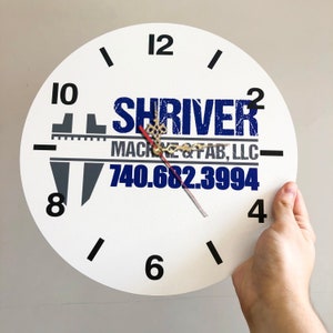 May include: White clock face with black numbers and hands. The clock has a blue and white logo that says "Shriver Machine & Fab, LLC" and a phone number "740.682.3994".