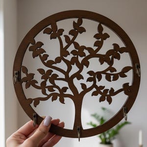 May include: A circular wooden key holder with a tree design. The dark brown key holder has a tree silhouette cut out of the centre. It features four metal hooks for hanging keys, and is made of wood.