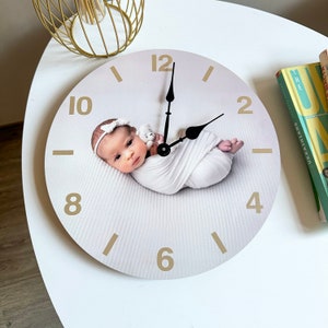 Puede incluir: Un reloj de pared redondo con una imagen de bebé. La esfera del reloj es de color beige claro con números dorados y manecillas negras. El bebé está envuelto en una manta blanca y lleva una diadema blanca. El reloj está sobre una superficie blanca.