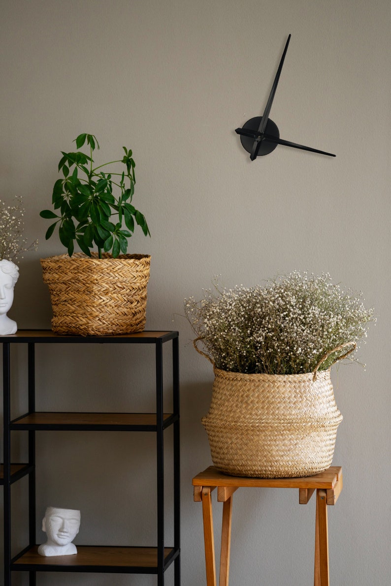 Op de afbeelding: Een zwarte klok met een ronde wijzerplaat en lange zwarte wijzers hangt aan een lichtgrijze muur. Twee rieten manden, een op een zwart metalen rek en de andere op een houten kruk, bevatten planten. De plant in de mand op het rek heeft groene bladeren, terwijl de plant in de mand op de kruk witte bloemen heeft.